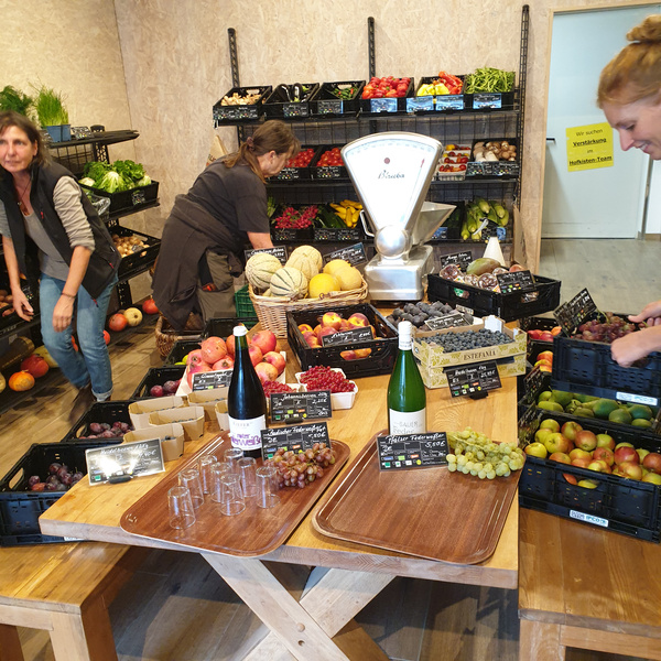 KI generiert: Ein Obst- und Gemüseladen mit Waage, Mitarbeitern und Produkten auf Tischen und Regalen. Text: „Wir suchen Verstärkung Hofladen-Team“.