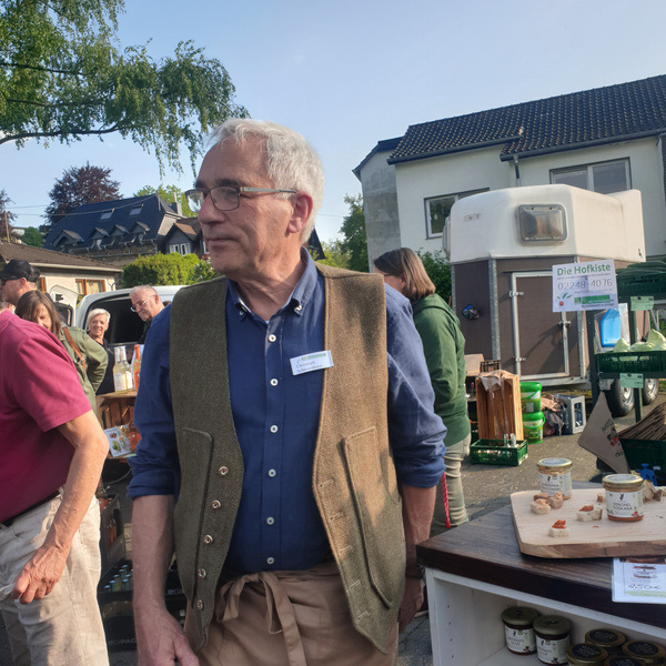 KI generiert: Ein Mann mit Weste steht vor einem Marktstand. Text: "Die Hofkiste 02243 4056".