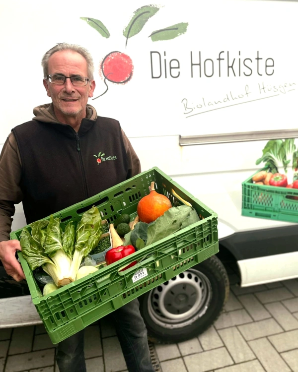 KI generiert: Ein Mann hält eine Kiste mit frischem Gemüse vor einem Lieferwagen. Text: "Die Hofkiste Biolandhof Hüsgen".