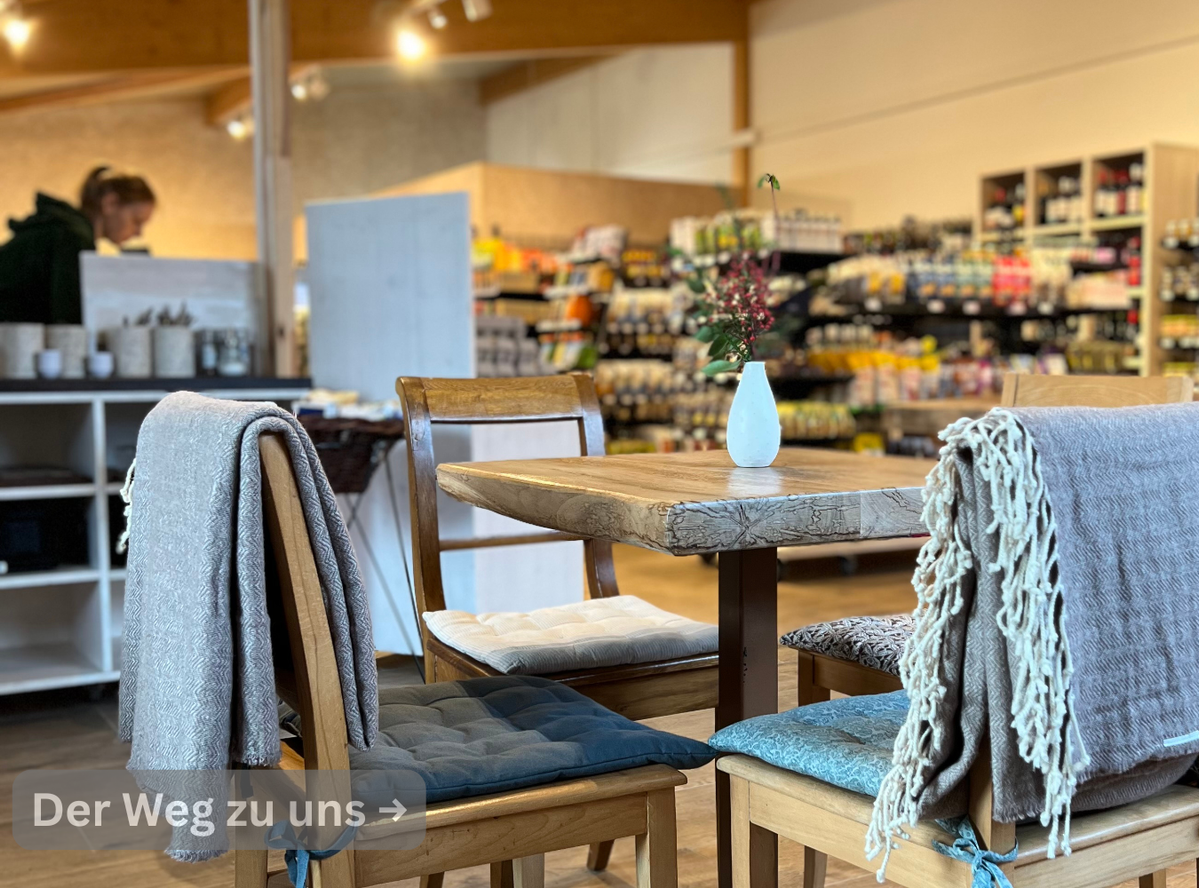 KI generiert: Gemütliches Café-Interieur mit Tisch und Stühlen, im Hintergrund ein Regal mit Produkten. Text: "Der Weg zu uns →".