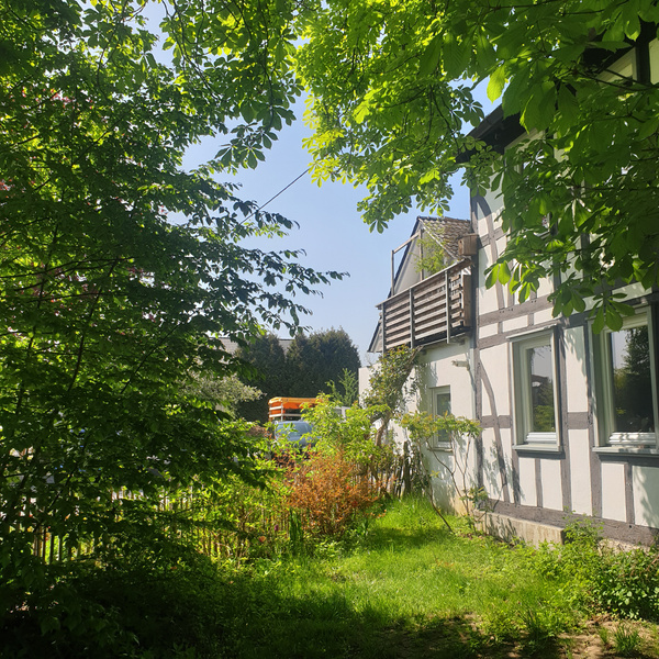 KI generiert: Ein Fachwerkhaus umgeben von grüner Vegetation an einem sonnigen Tag.