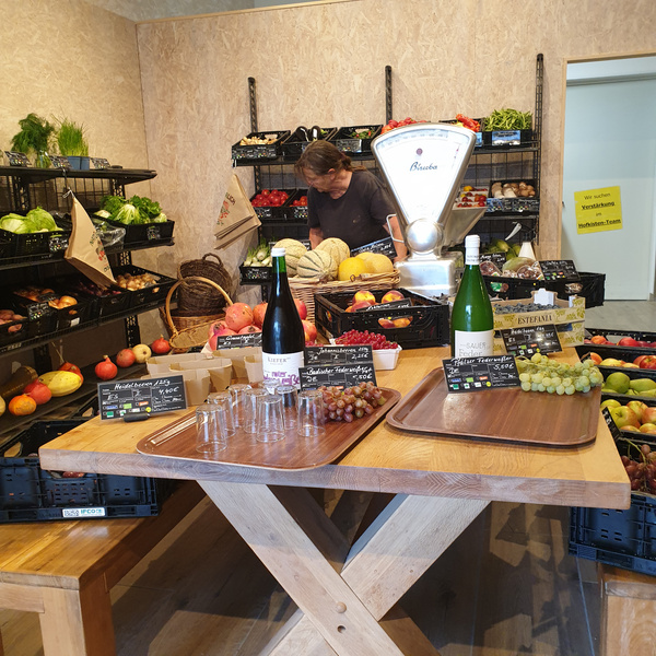 KI generiert: Ein kleiner Obst- und Gemüseladen mit Weinflaschen und Verkostungsproben. Text: "Zur Verstärkung unseres Hofladen-Teams!"