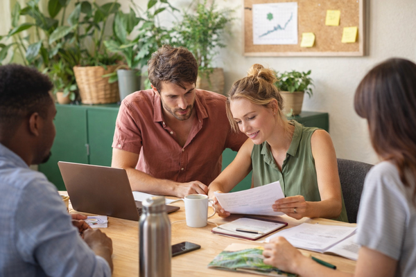 KI generiert: Gruppe im Büro beim Besprechen von Dokumenten, umgeben von Pflanzen, auf Wand eine Pinnwand mit Grafiken.