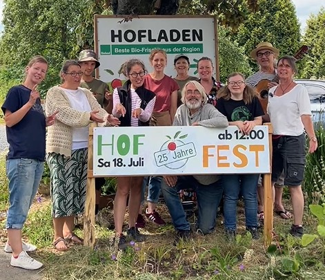 KI generiert: Eine Gruppe von Menschen posiert vor einem Schild mit der Aufschrift: "HOFLADEN", "HOF FEST Sa 18. Juli ab 12:00", "25 Jahre".